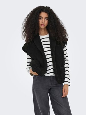 Jeune femme aux cheveux bouclés portant un gilet en polaire noir par-dessus un t-shirt à manches longues blanc et noir rayé et un jean foncé sur un fond uni.