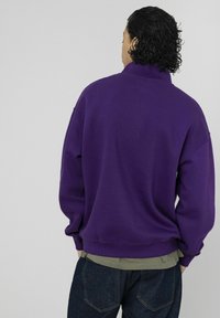 Sweat-shirt violet à col haut et épaules tombantes, associé à un sous-pull vert et un jean bleu foncé. Texture lisse, coupe décontractée.