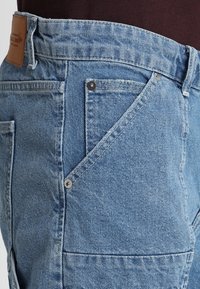 Ljusblå denimjeans med en klassisk design, synliga sömmar och en distinkt vinklad ficka med en metallknappdetalj.