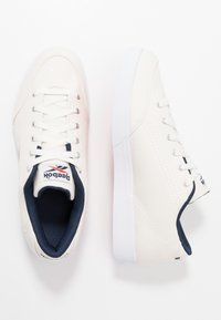 Zapatillas de lona blancas con detalles en marino, cordones planos, puntera redonda y suela de goma texturizada. Presentan el logo de Reebok en la lengüeta y en el talón.