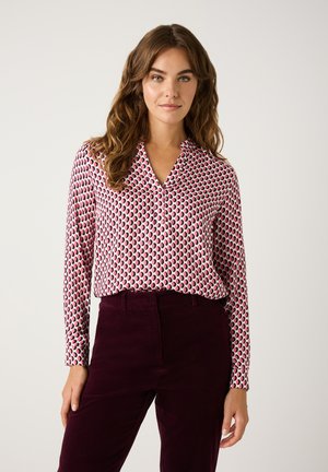 Long-sleeve blouse in white with a red and black circular pattern. Soft fabric, collarless V-neck, paired with dark burgundy pants.