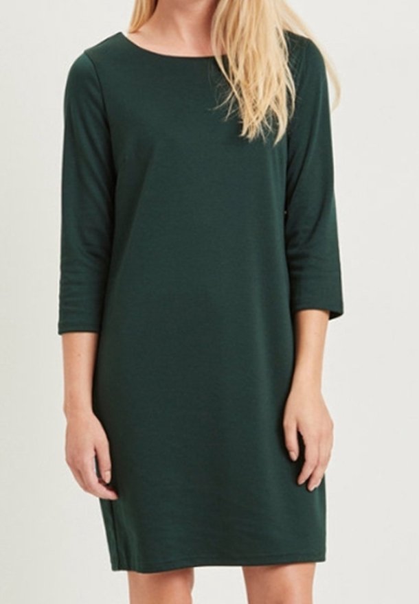 Femme portant une robe vert foncé longueur genou avec des manches trois-quarts et un col rond, debout devant un fond uni.