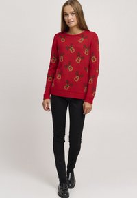 Pull rouge avec un motif floral en jaune et vert, manches longues, col rond, ourlet côtelé, associé à un pantalon et des chaussures noirs.
