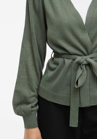 Cardigan de punto verde oliva con un profundo escote en V, cintura ajustable, mangas abullonadas y detalles de patrón sutiles en toda la prenda. Textura suave y forma ajustada.