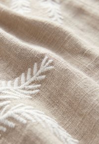 Beige stof met witte geborduurde bladpatronen; zachte textuur met een geweven uiterlijk, met subtiele variaties in weefsel en ontwerp.