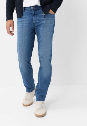 Mann trägt blaue Jeans, weiße Sneaker und eine dunkle Jacke mit einer Hand in der Tasche, steht vor einem schlichten weißen Hintergrund.