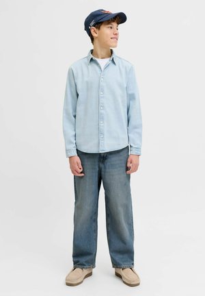 Nastolatek w granatowej czapce, jasnoniebieskiej koszuli na guziki, szerokich dżinsach i beżowych butach, stojący i patrzący w prawo.