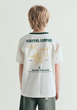 Kind met blond haar, draagt een wit T-shirt met een groene kraag en een citroenthema-print en tekst op de achterkant.