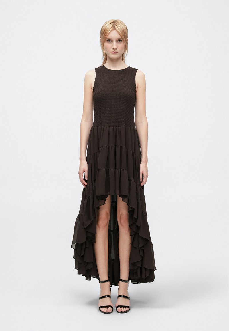 Nő áll sleeveless fekete high-low ruhában, fodros szegéllyel és fekete pántos magassarkúban, sima fehér háttér előtt.