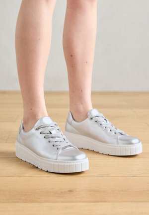 Persoon die zilverkleurige metallic vetersneakers met dikke witte zolen draagt en op een lichte houten vloer staat.