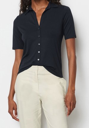 Kvinde iført en sort kortærmet skjorte med knapper, let stoppet ned i cremefarvede bukser mod en ensfarvet hvid baggrund.