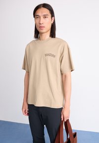 Beige cotton t-shirt with short sleeves and a round neckline. Features a small black "DICKIES" logo on the left chest.