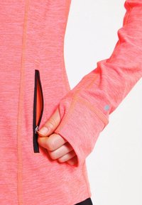 Hånd, der trækker lynlås på frontlomme af pink langærmet træningstrøje med tommelfingerhul og reflekterende prik på ærmet.