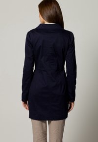 Laivastonsininen trenssitakki, jossa on kaulus, istutettu vyötärö ja pitkät hihat. Valmistettu sileästä kankaasta, pituus puoliväliin reittä ja takana saumat.