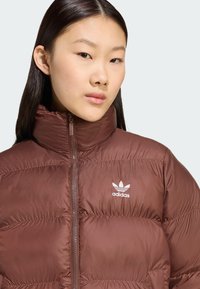 Brun puffer-jakke med høj krave, lynlåslukning og hvidt Adidas-logo. Glat tekstur med kviltet mønster og polstrede sektioner.
