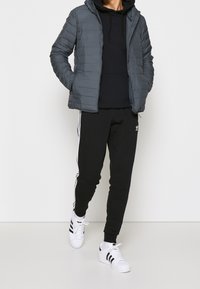 Giacca piumino grigia con cappuccio, felpa nera sotto, pantaloni sportivi neri con strisce bianche e sneakers bianche con dettagli neri.