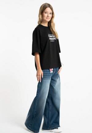 Młoda kobieta ubrana w czarny t-shirt z nadrukiem i szerokie niebieskie dżinsy, stoi na białym tle, lekko się uśmiecha, ręce swobodnie opuszczone wzdłuż ciała.