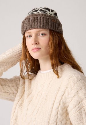 Bruine gebreide beanie met een wit patroon bovenop, geribbelde rand en een klein leren label. Model draagt een crèmekleurige kabelgebreide trui.