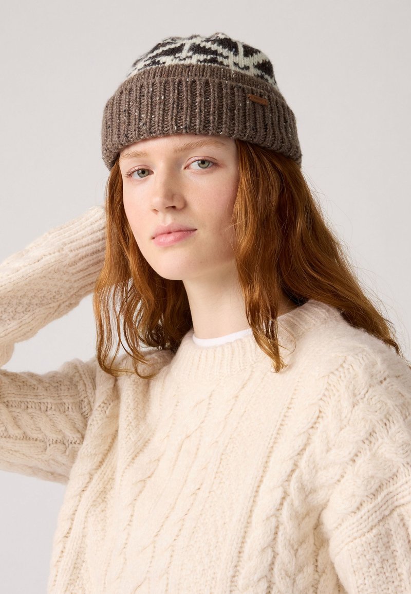 Brown knitted beanie with white pattern on top, ribbed band, and a small leather tag. Model wears a cream cable-knit sweater.