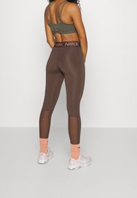 Hnědé sportovní legíny s mřížkovými panely, vysokým pasem a logem Nike, doplněné zelenou sportovní podprsenkou a růžovými ponožkami.