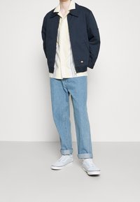 Persona che indossa una giacca bomber blu navy, una camicia a righe giallo chiaro, jeans azzurri risvoltati e sneakers bianche, in piedi contro uno sfondo neutro.