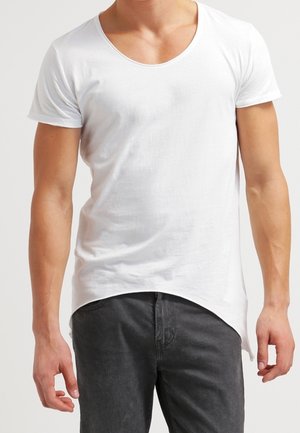 Mężczyzna noszący dopasowany biały T-shirt z krótkim rękawem i zaokrąglonym dołem, w połączeniu z ciemnoszarymi spodniami, stojący na jednolitym tle.