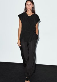 Top nero senza maniche con scollo drappeggiato, abbinato a pantaloni neri larghi. L'outfit presenta una vita cinturata e un tessuto morbido.