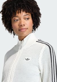 Bijela jakna s patentnim zatvaračem i visokim ovratnikom, s crnim trostrukim prugama na ramenima i izvezenim logotipom na prsima.