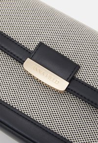 Borsa nera e beige caratterizzata da un tessuto testurizzato con piccoli motivi perforati, una tracolla in pelle e un accento in metallo dorato con il logo.