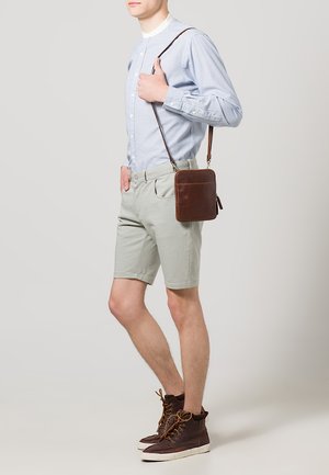 Braune Leder-Umhängetasche, hellblaues gestreiftes Hemd mit Knopf, hellgraue Shorts, braune Schnürstiefel. Lässige, figurbetonte Silhouette.