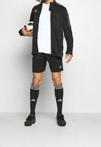 Sort atletisk jakke med regnbuefarvede stribe, hvid t-shirt, sorte shorts, knælangen sorte sokker med grå striber, og sorte fodboldstøvler.