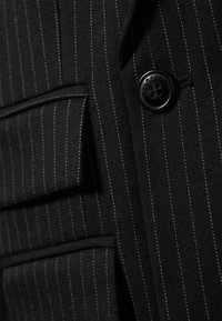 Svart stripete stoff har en enkelt knappelukking og en dekorativ klafflomme, med kontrasterende hvite sømmer.