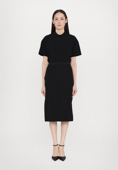 Schwarzes Zweiteiler-Outfit bestehend aus einem kurzärmeligen Oberteil und einem knielangen Rock. Glatte Textur, minimalistisches Design, kombiniert mit schwarzen spitzen Absätzen.