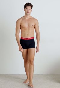 Schwarze Boxerbriefs mit rotem Bund, der das "TOMMY HILFIGER"-Logo trägt. Das Modell steht barfuß vor einem neutralen Hintergrund.