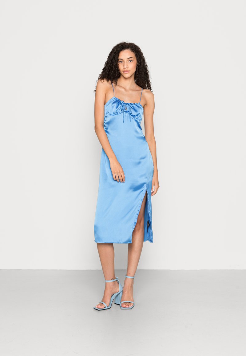 Vestido midi de cetim em azul claro com alças finas ajustáveis, corpete franzido, abertura lateral e textura suave. Usado com sandálias de salto azuis.