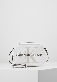 Borsa a tracolla bianca realizzata in materiale liscio, con un grande logo "CK" e la scritta "CALVIN KLEIN JEANS" stampata in grassetto nero. Tracolla con dettagli neri.