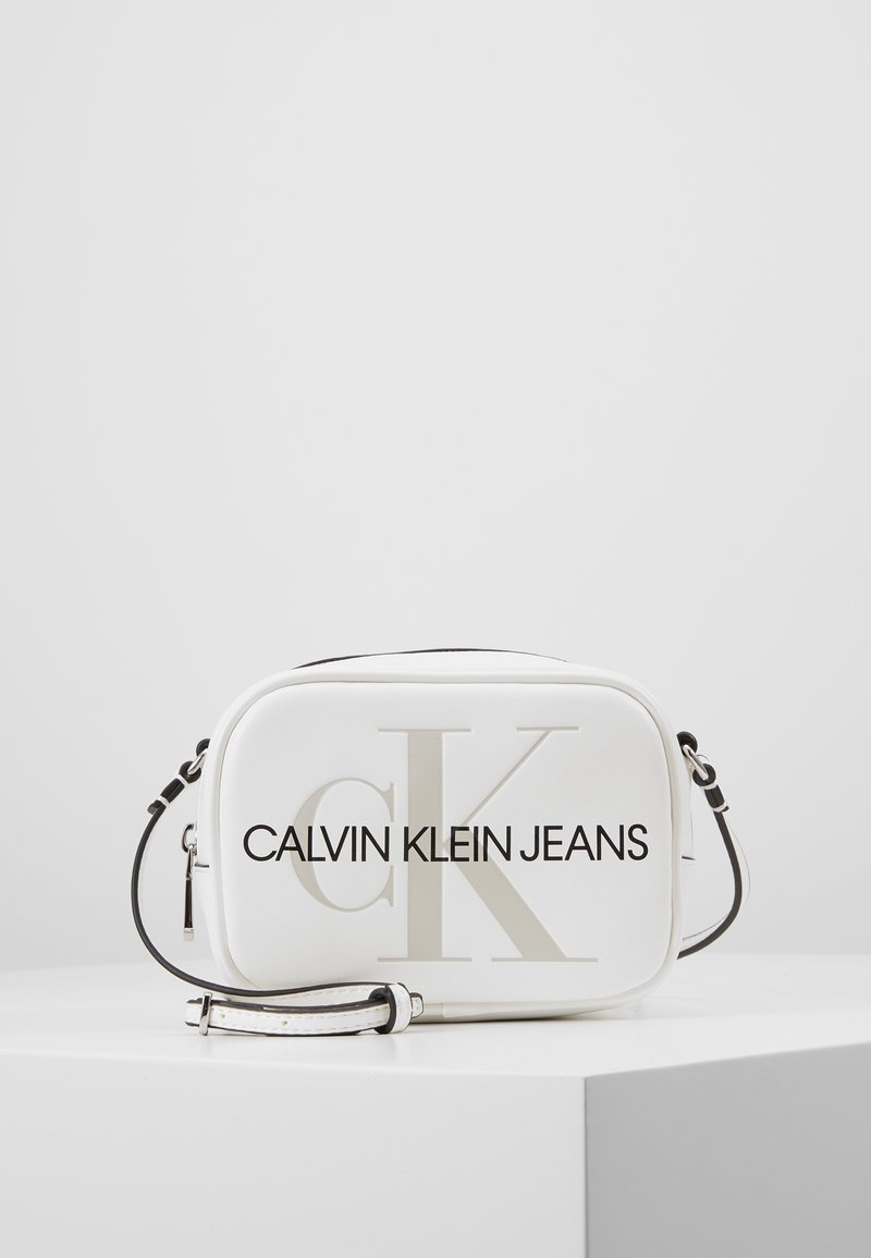 Borsa a tracolla bianca realizzata in materiale liscio, con un grande logo "CK" e la scritta "CALVIN KLEIN JEANS" stampata in grassetto nero. Tracolla con dettagli neri.