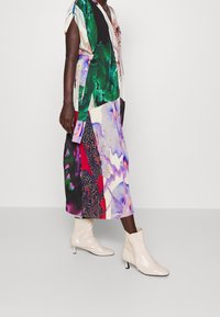 Robe multicolore avec un mélange de motifs abstraits, manches courtes, et taille nouée, associée à des bottines beiges à la cheville et un talon pointu.
