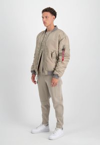 Chaqueta bomber beige con cierre de cremallera, forro acolchado y una etiqueta roja en la manga, combinada con pantalones de chándal beige ajustados y zapatillas blancas.