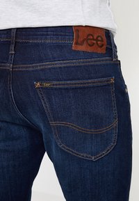 Jean en denim bleu foncé avec surpiqûres orange, une poche arrière et un patch en cuir marron de la marque Lee sur la ceinture.