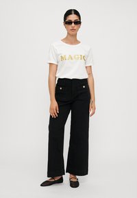 White t-shirt with gold glitter "MAGIC" text, paired with high-waisted black wide-leg pants. Model wears black flat shoes and sunglasses.