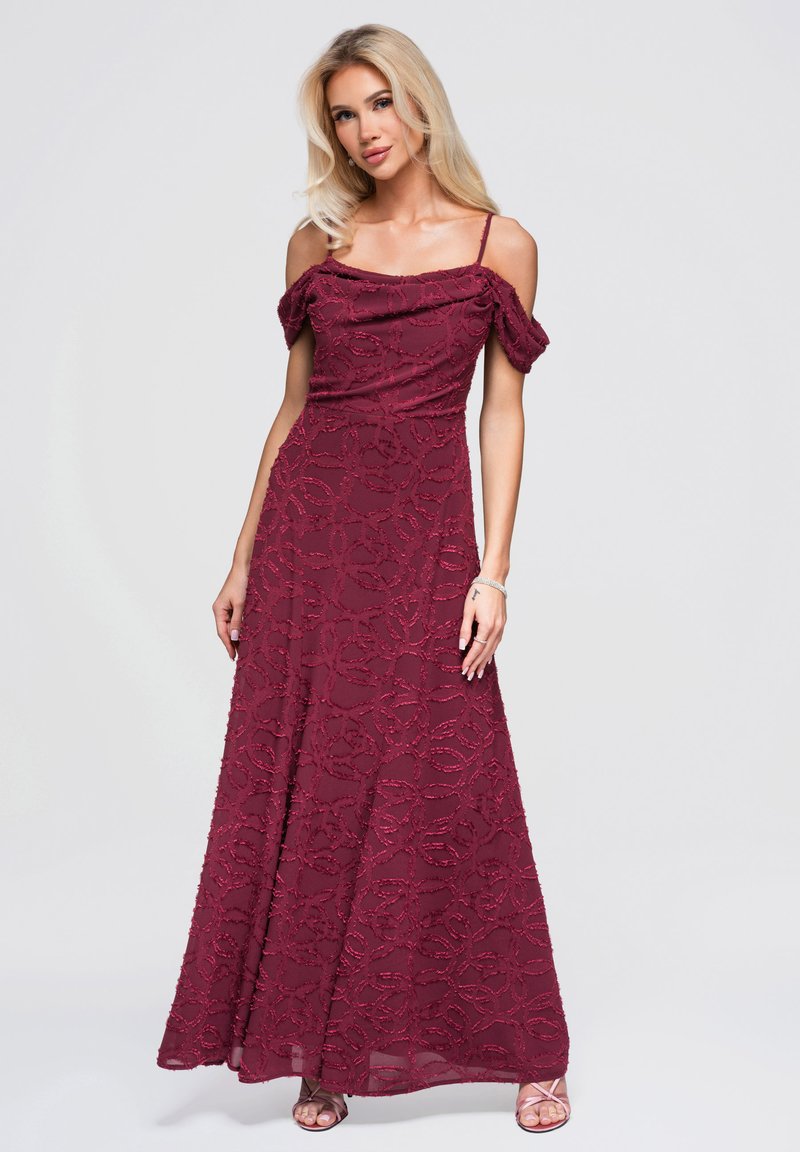 Femme aux longs cheveux blonds portant une robe bordeaux longue jusqu'au sol, épaules dénudées, avec des motifs texturés et des talons ouverts, debout devant un fond uni.