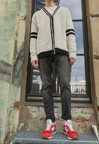 Bílý kardigan s námořnickými pruhy, knoflíky vpředu, kombinovaný s tmavě modrými džíny a červenými teniskami New Balance s multibarevnými akcenty.