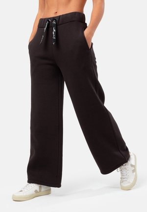 Person trägt lockere schwarze Sweatpants mit Kordelzug und weiße Turnschuhe, steht mit den Händen in den Taschen.