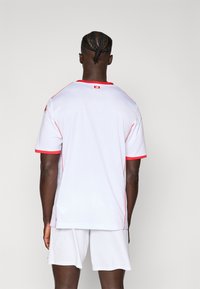 Balta sporta krekla ar sarkaniem akcentiem, īsām piedurknēm un apaļu apkakli. Gluda tekstūra, uz aizmugures netālu no kakla ir logotips.