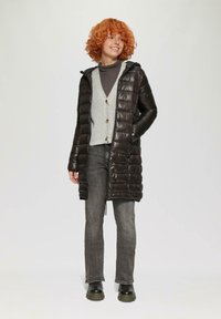 Zwarte pufferjas met capuchon, voorzien van een gewatteerd ontwerp en glanzende textuur. Draag deze over een grijze cardigan, gecombineerd met grijze jeans en zwarte laarzen.