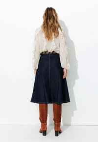 Jupe en denim évasée avec un ourlet brut, portée avec un blouse crème à volants. Associe à des bottes à talons haute et en suède marron, arrivant jusqu'aux genoux.