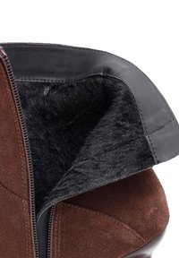 Stivale in suede marrone con rifiniture in pelle nera e una morbida fodera nera. Presenta una chiusura con zip lungo il lato. La parte interna è soffice e strutturata.