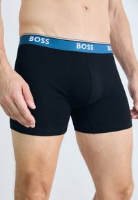 Black boxer trunks with a soft waistband featuring blue and white "BOSS" logo. Made from stretch cotton, form-fitting design, smooth texture.