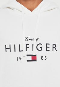 Hvit hettegenser med en svart "Tommy Hilfiger"-logo og et rødt og blått rektangulært emblem, samt datoen "1985" trykket under. Myk stofftekstur.